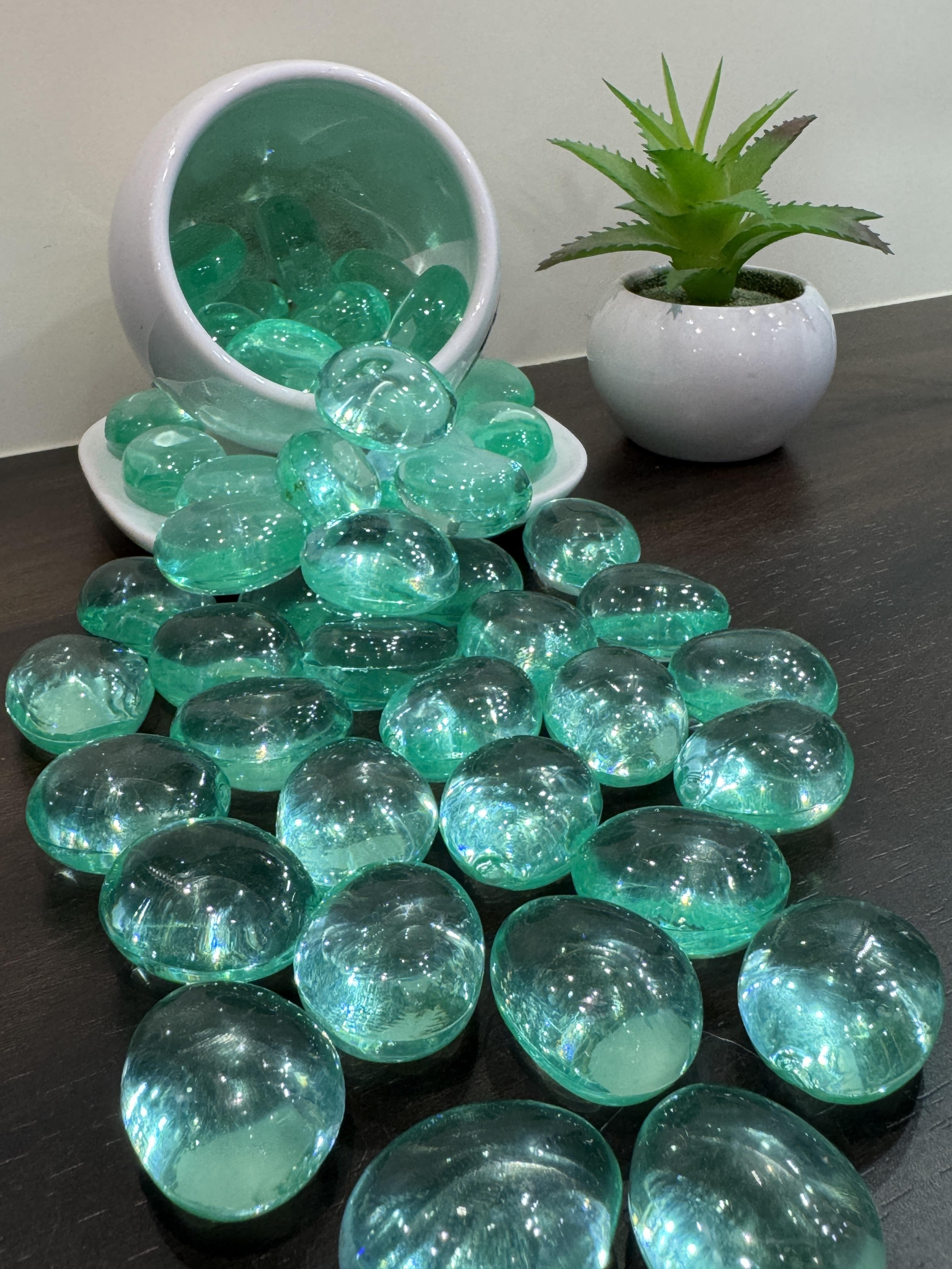 Teal glass marbles on a dark surface with a white bowl and potted plant in the background.