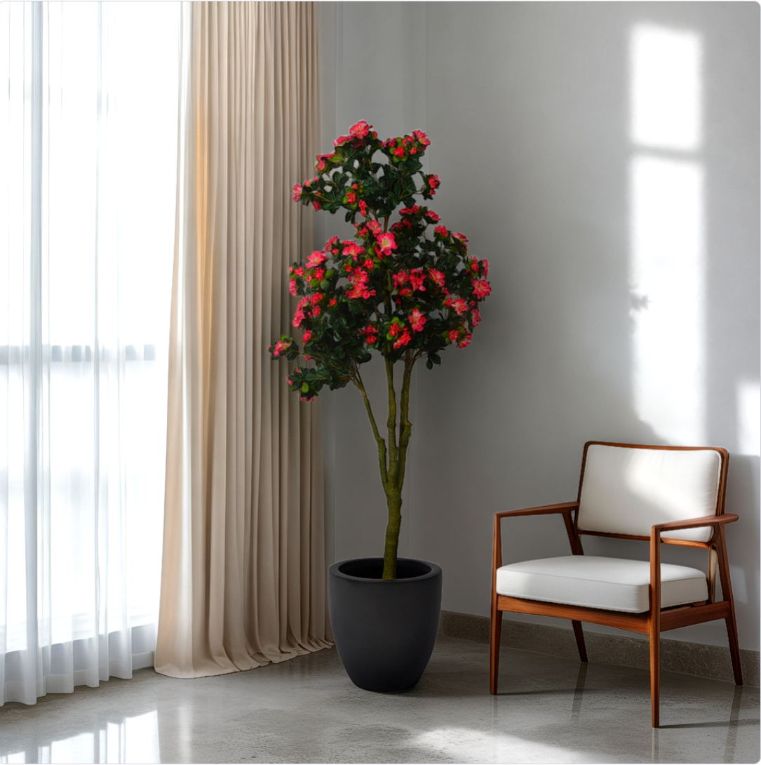 A 5 ft artificial azalea flower plant with green and red leaves in a room corner next to a chair, placed in a black pot.