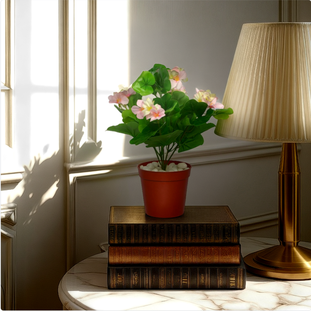 An artificial green geranium bush in a red pot, placed on a stack of books beside a lamp, with sunlight streaming through a window.