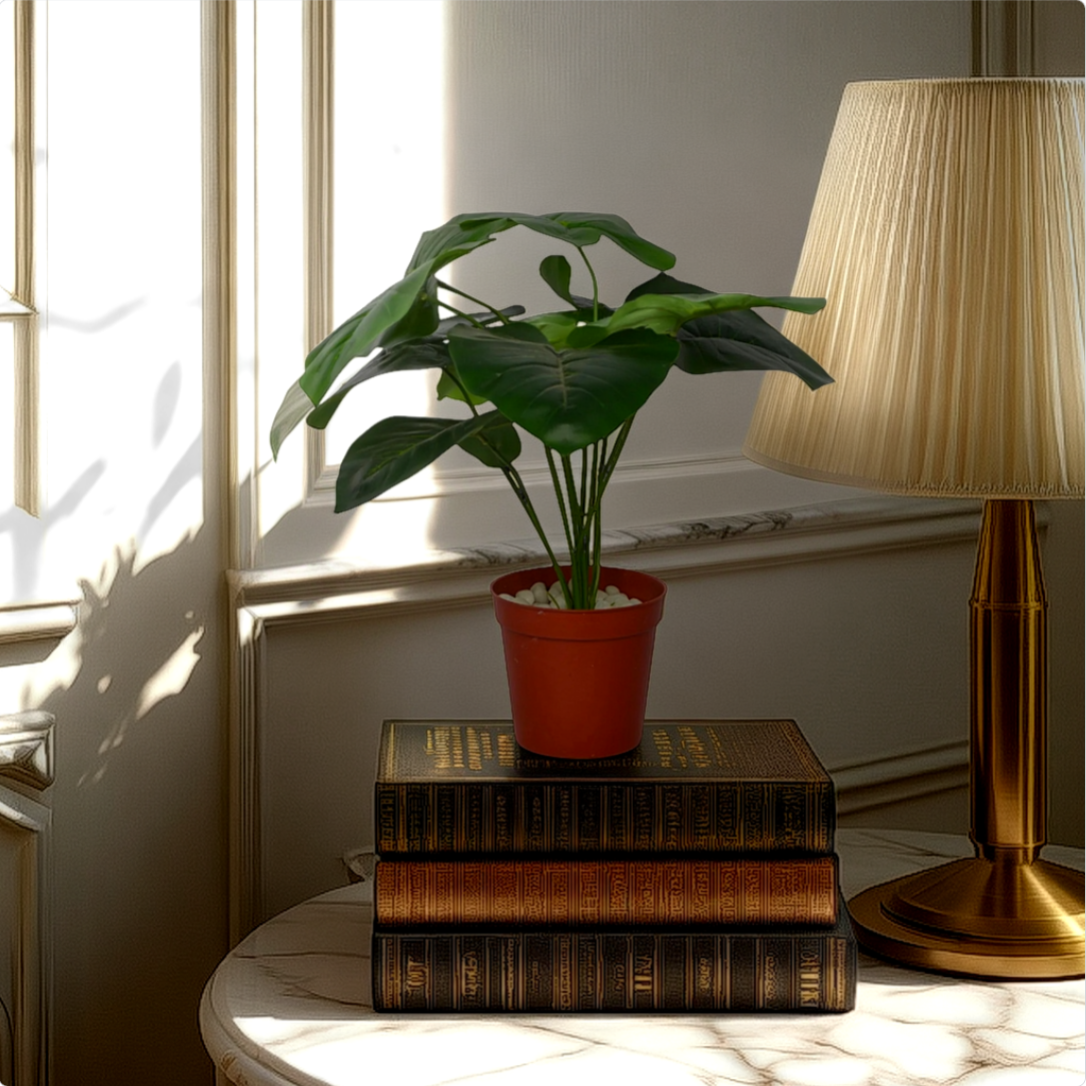 An artificial Alocasia bush in a red pot, placed on a stack of vintage books on a side table, with a lamp and window light in the background.