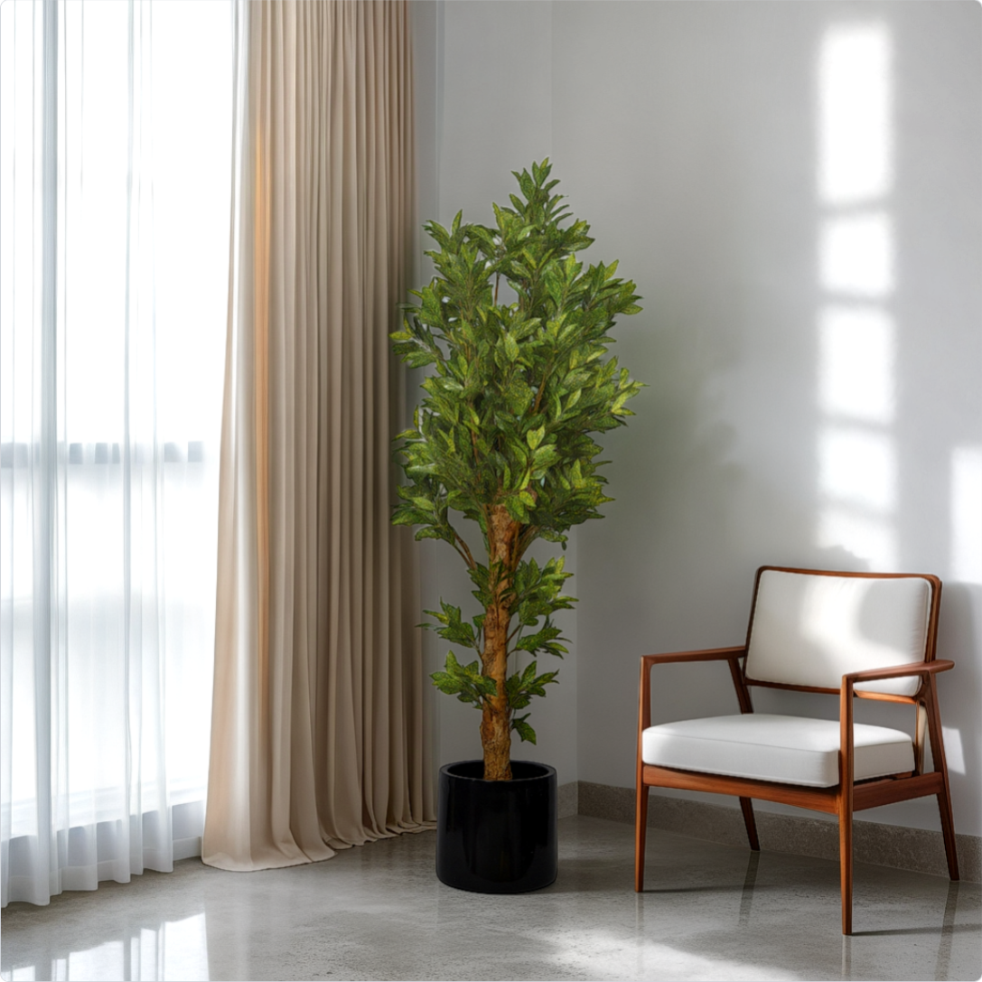 A 5 ft artificial croton plant with multicolor leaves placed in a room corner beside a window with curtains, next to a chair with a white cushion.