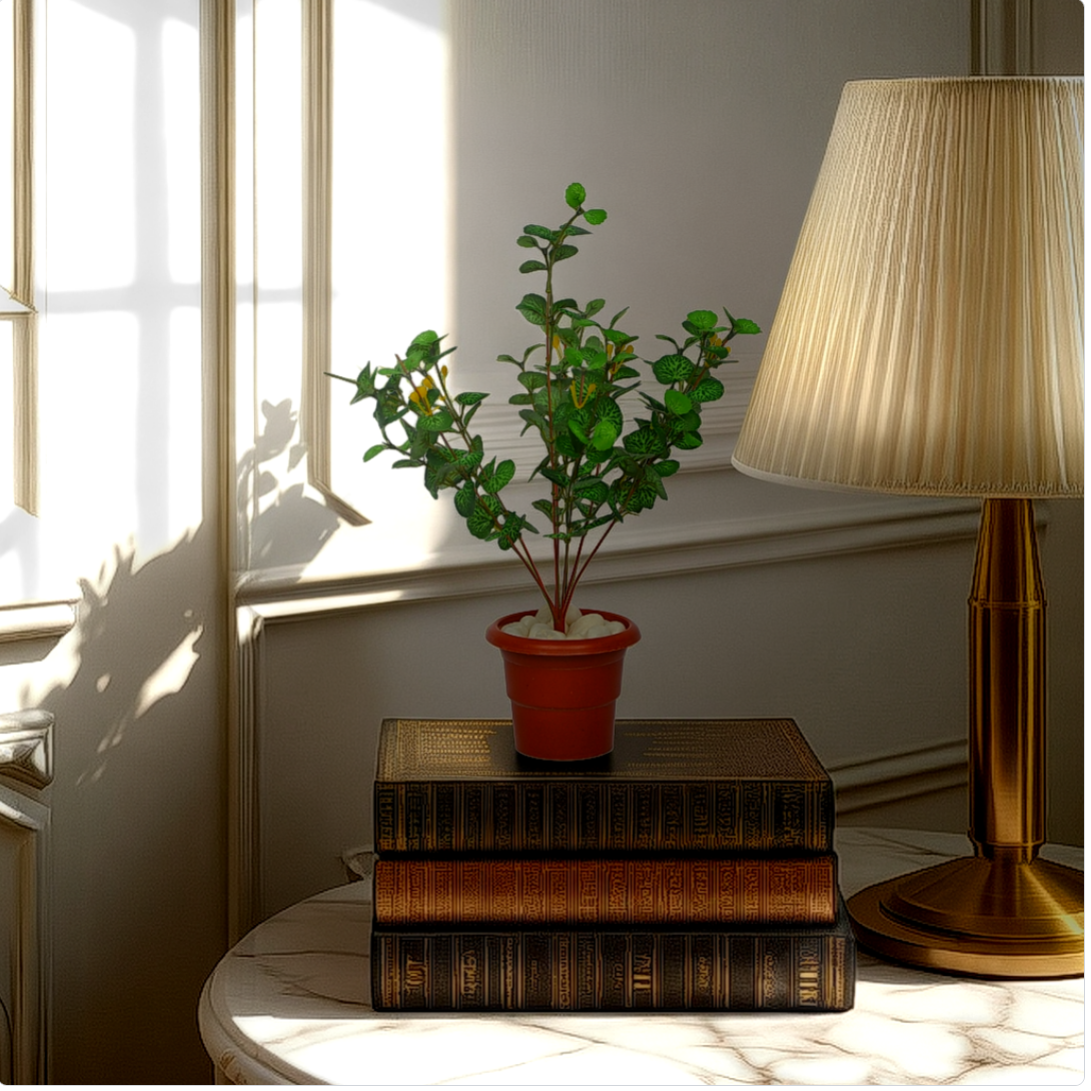 An artificial Geranium plant in a red pot, placed on a stack of old books, with a lamp and a window in the background.