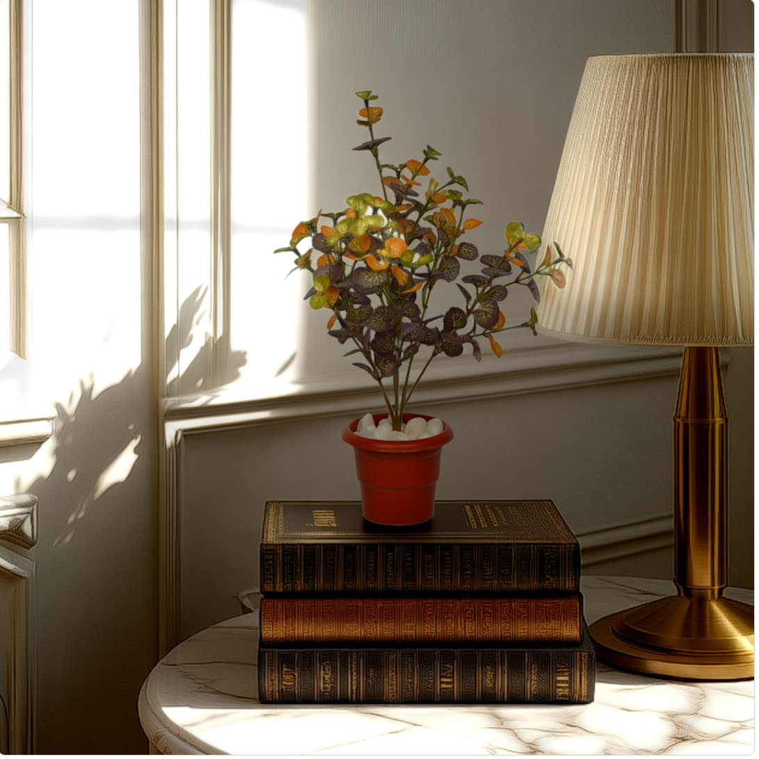 An artificial geranium plant in a red pot, placed on a stack of books, with a lamp and a window in the background.