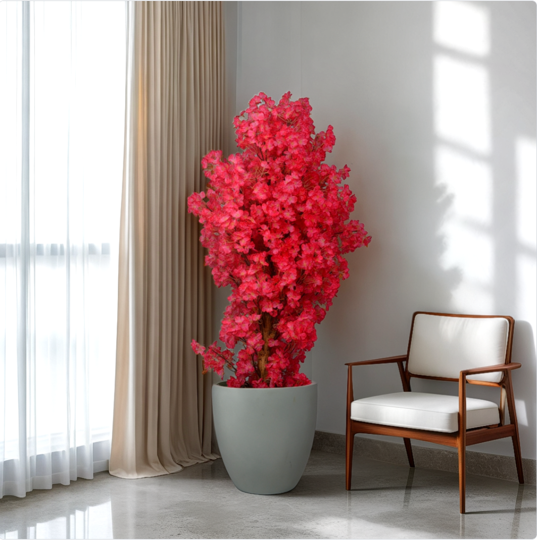 A 5 ft artificial cherry blossom plant with red flowers in a grey pot, placed in a home interior setting next to a window with light coming through and a white chair.