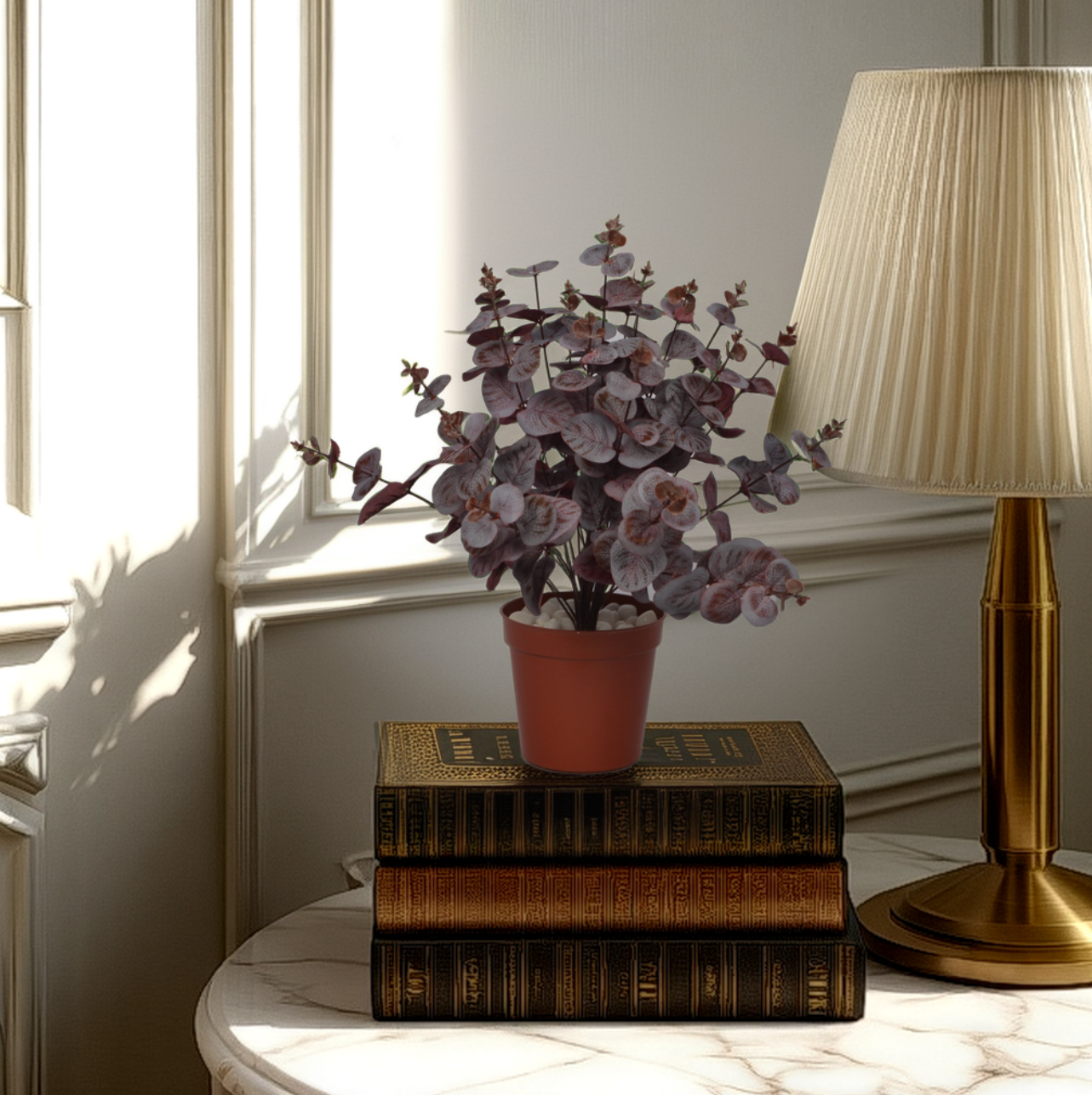 An artificial carpinteria bush with red leaves, placed on top of a stack of books, next to a lamp with a beige lampshade, on a white surface.