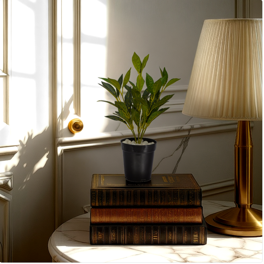 An artificial yellow mini croton bush in a black pot placed on a stack of brown books, next to a lamp, in a room with natural light coming through the window.