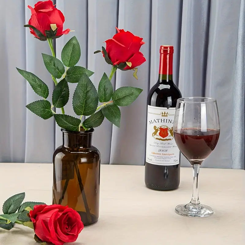 Red roses in a vase, bottle of wine, and glass on a table with a gray curtain background