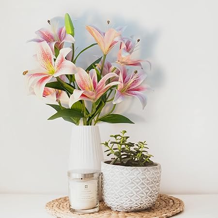 Bouquet of pink lilies in a white vase with a small potted plant and candle on a woven coaster.