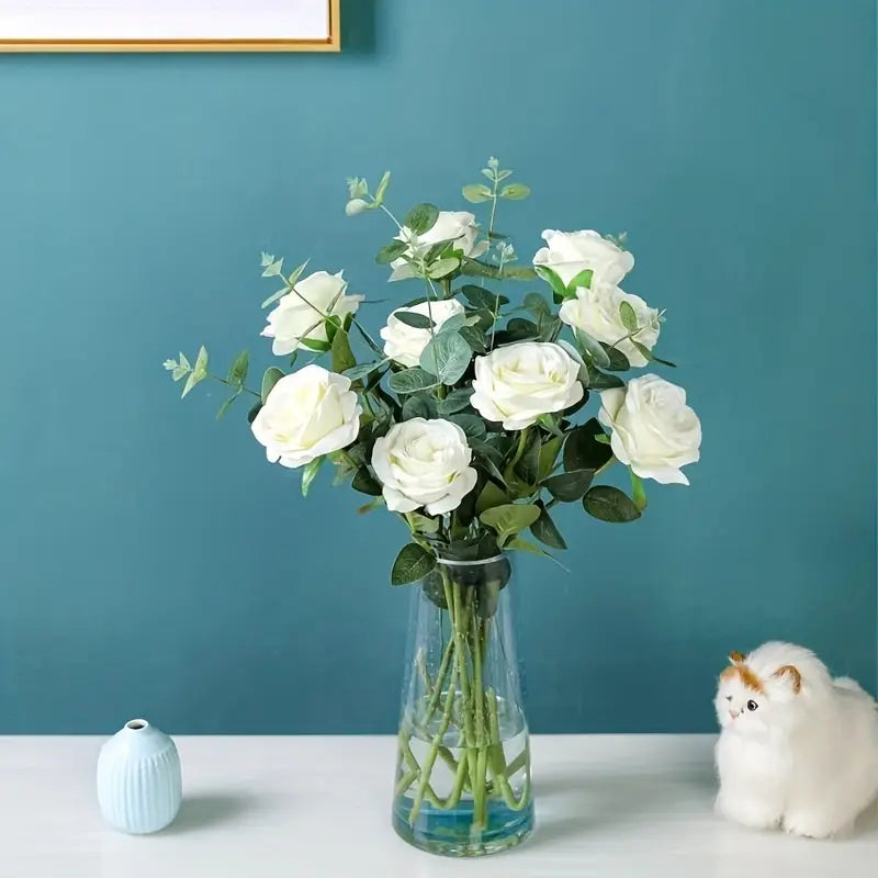 Bouquet of white roses in a clear vase against a teal wall with a small white dog figurine.