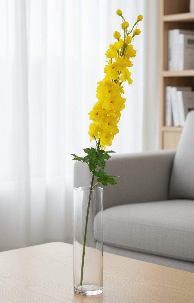 Yellow flowers in a clear vase on a wooden table with a gray sofa and bookshelf in the background.