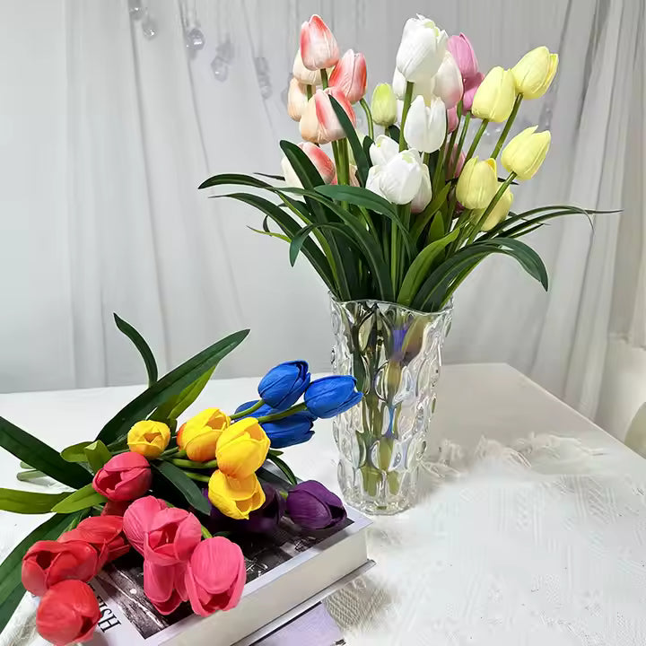 Bouquets of colorful tulips in a clear vase on a white surface with a light curtain background.