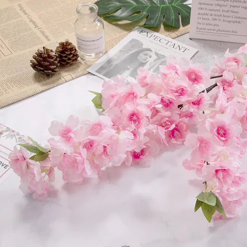 Pink artificial flowers on a newspaper with a bottle and pinecones in the background