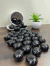 Black stones on a wooden surface with a small white bowl and green plant in the background.