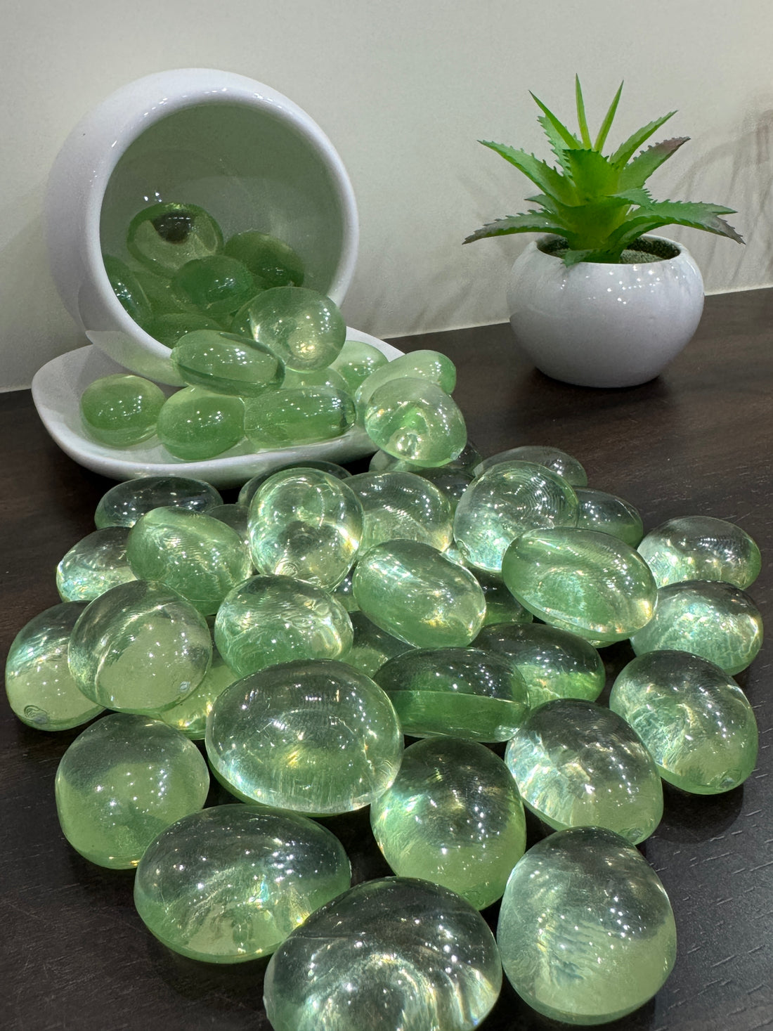Green glass pebbles on a dark surface with a white bowl and plant in the background.