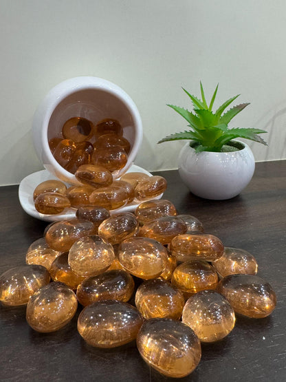 Amber glass beads on a dark surface with a white bowl and plant in the background.