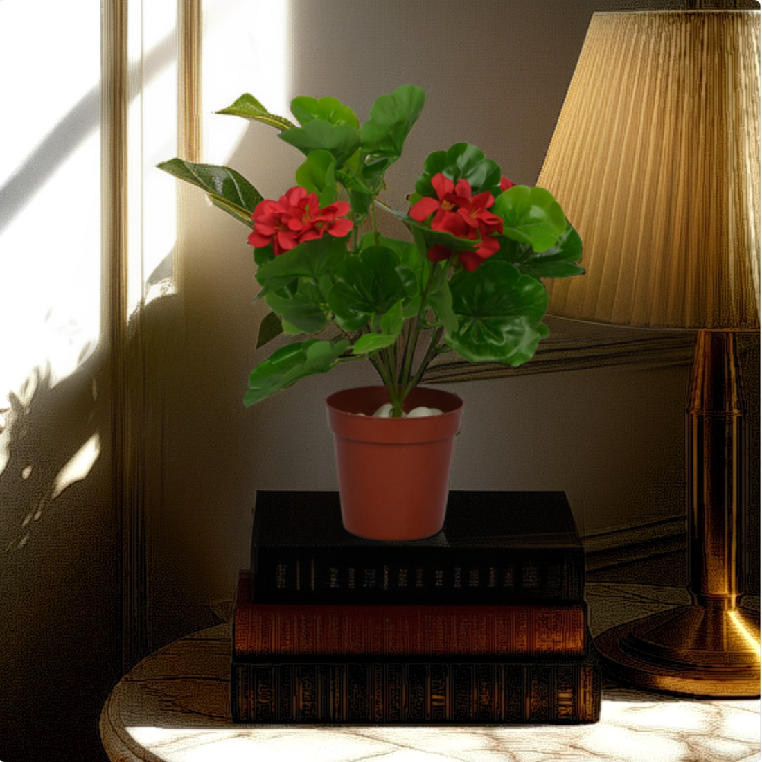 An artificial geranium plant in a brown pot, placed on a stack of books beside a lamp, with a realistic appearance.