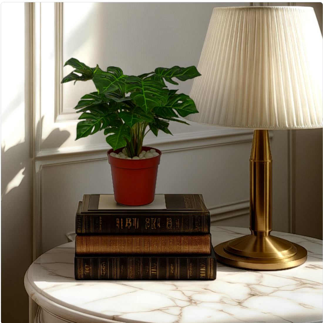 An artificial Monstera plant in a red pot, placed on top of a stack of vintage books, next to a classic style lamp with a beige lampshade, all placed on a white surface under natural lighting.