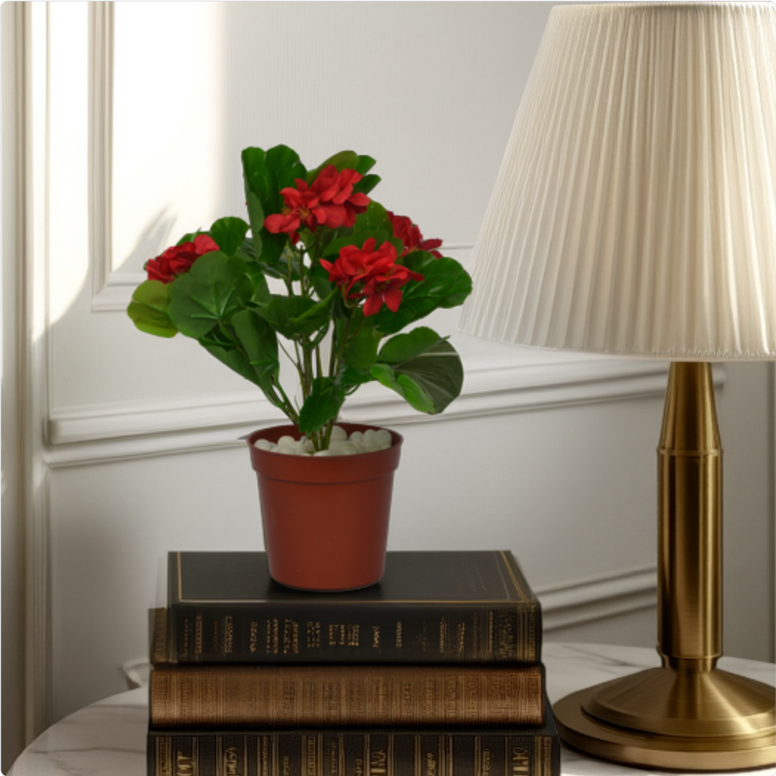 An artificial red geranium bush in a brown pot placed on a stack of books next to a lamp.