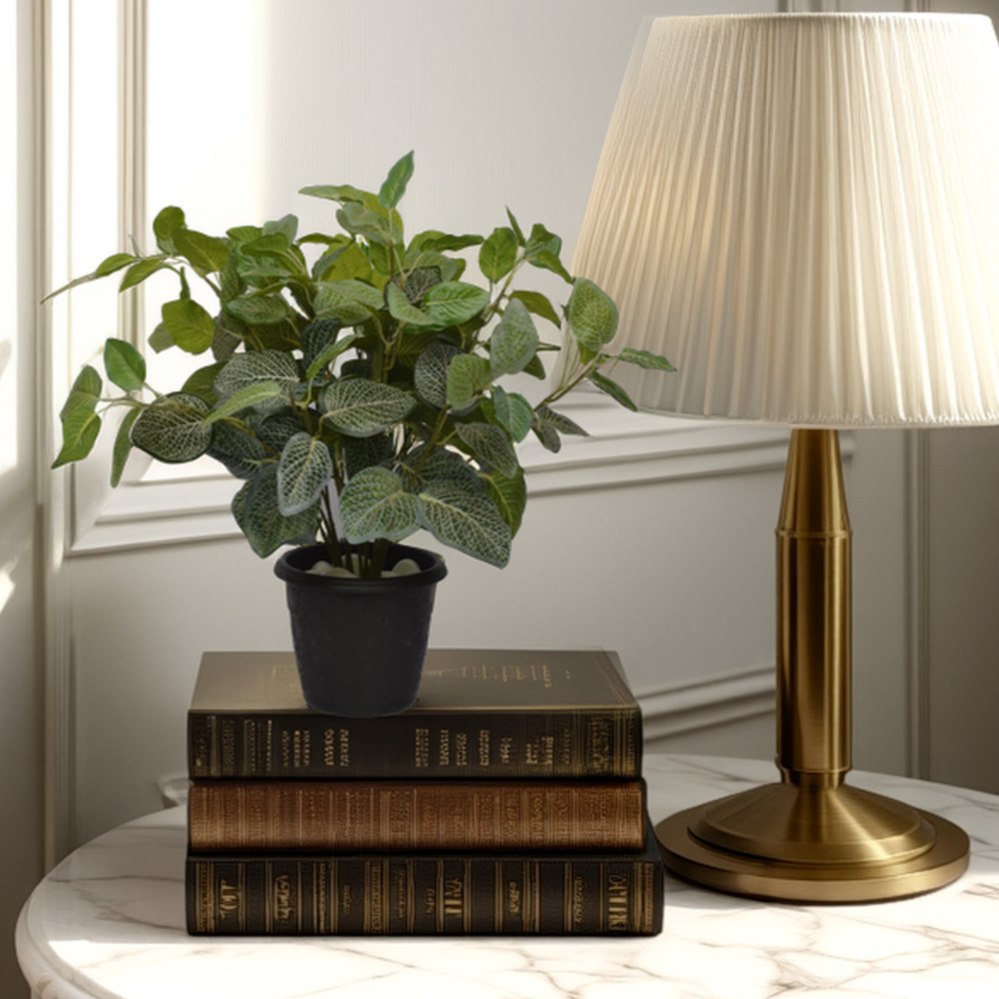 An artificial Fittonia bush in a black pot, placed on a stack of brown books next to a brass lamp with a white shade, on a white surface with a decorative molding in the background.
