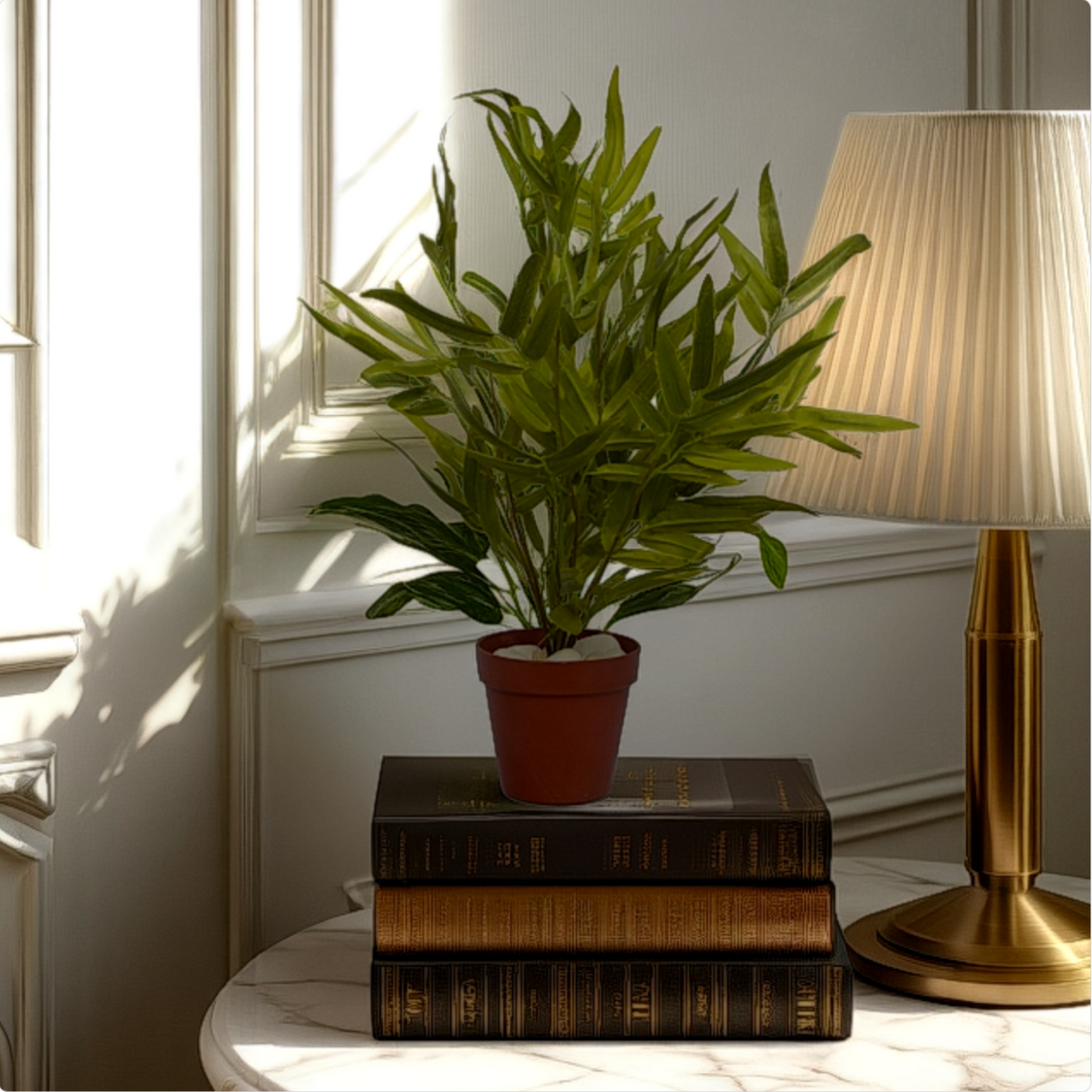 An artificial bamboo leaf bush in a red pot, placed on a stack of vintage books next to a lamp, indoors.
