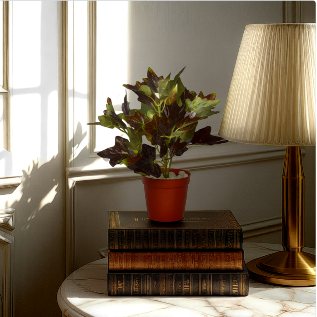 An artificial mini bush in a red pot placed on a stack of books, next to a lamp, with a sunlit background.
