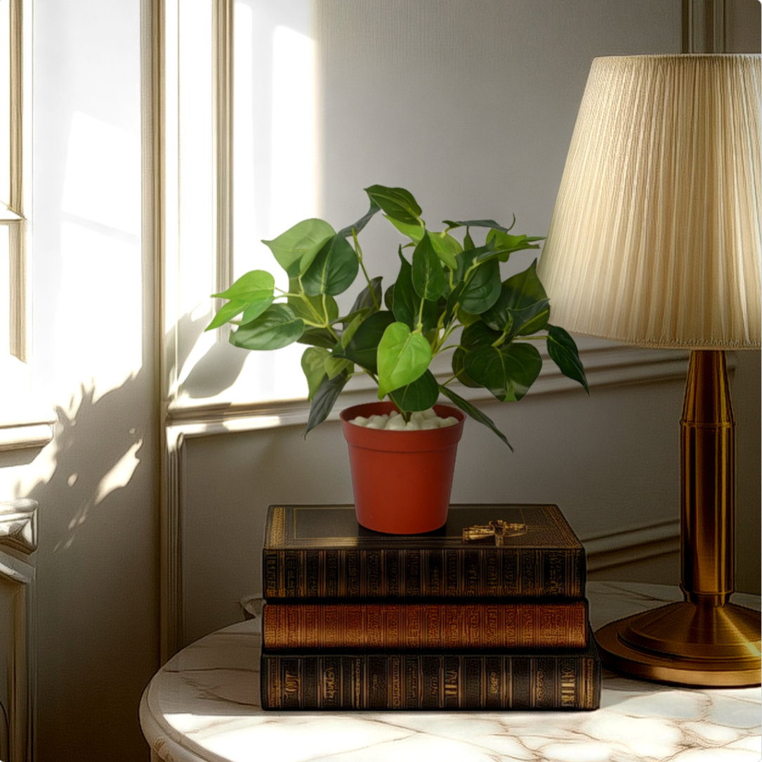 An artificial green betel bush plant in a red pot, placed on a stack of books on a table, with a lamp and window light providing illumination.