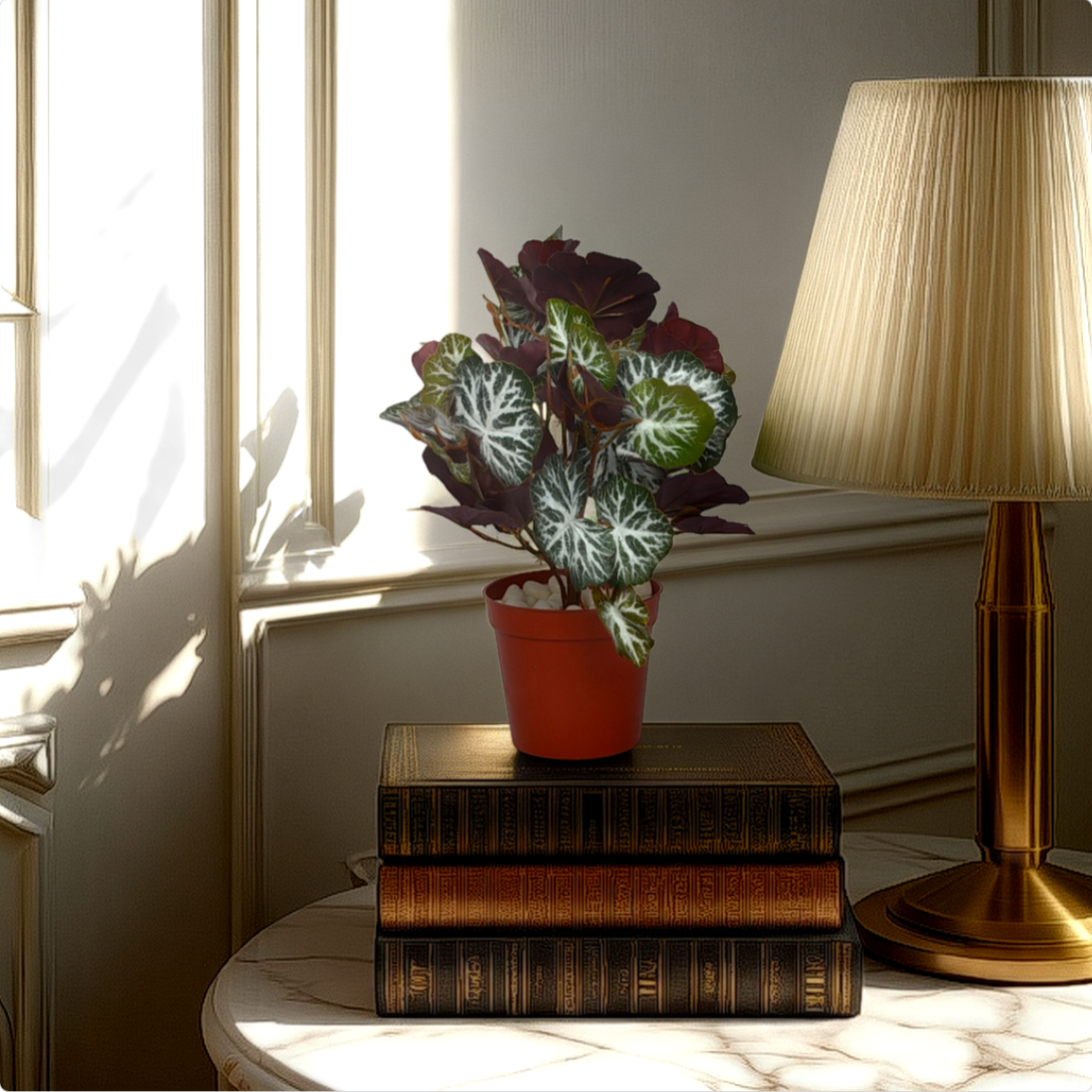 An artificial begonia bush in a red pot placed on a stack of books, next to a lamp, with a window in the background.