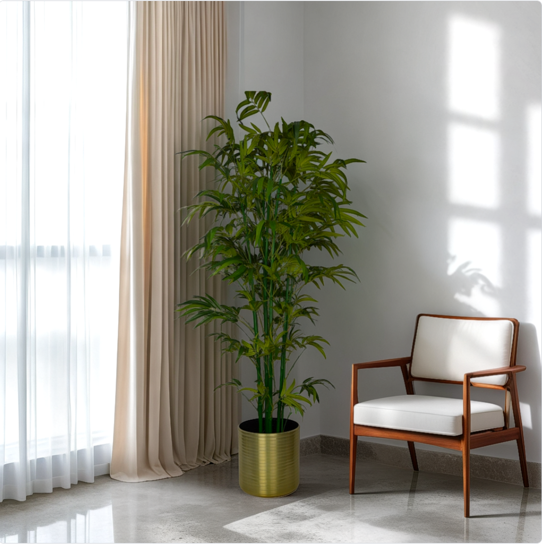 A 6 foot artificial bamboo tree in a gold pot placed in a corner of a room with a chair and a window in the background.