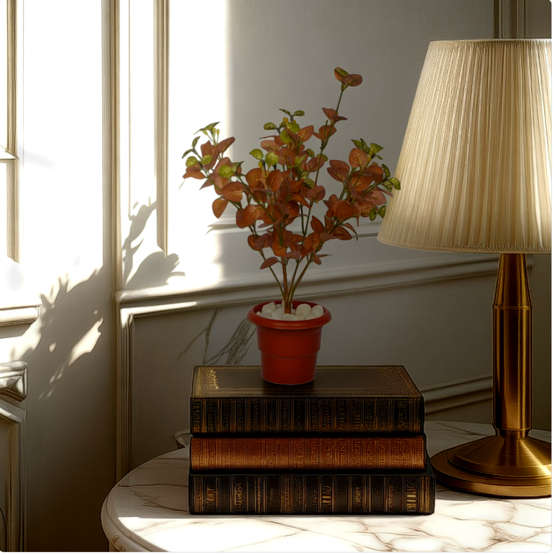 An artificial green multicolor carpinteria bush in a red pot, placed on a stack of old books, next to a classic style lamp, with sunlight streaming through a window.