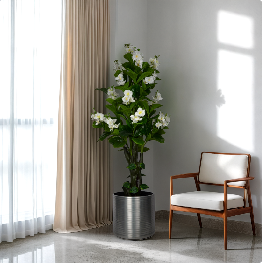 A 6 ft artificial camellia plant in a silver pot placed in a room corner beside a window with curtains. A chair with a white cushion is also visible in the image.