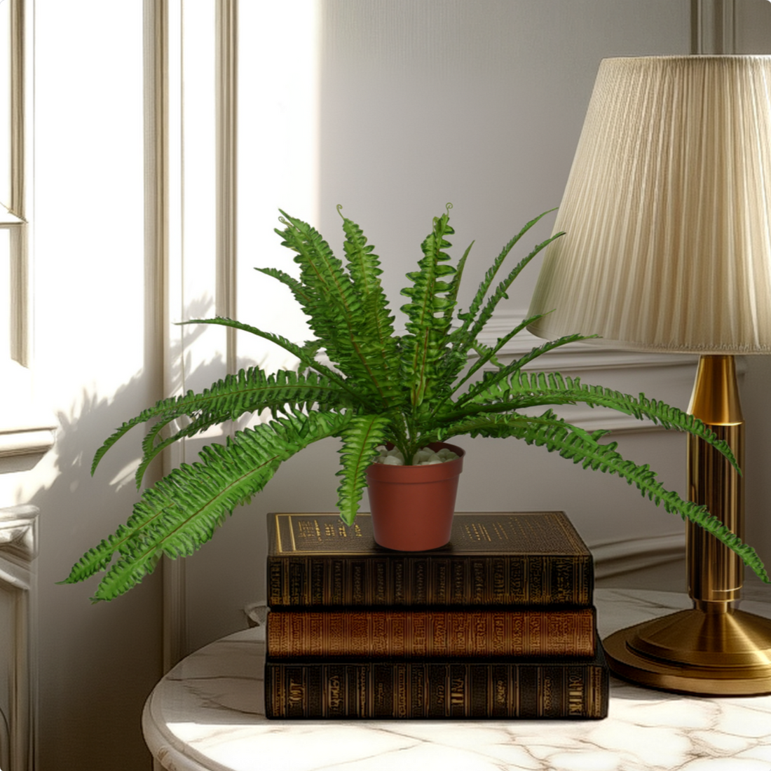 An artificial green fern bush in a brown pot, placed on top of a stack of books, next to a lamp on a marble surface, with a window in the background.