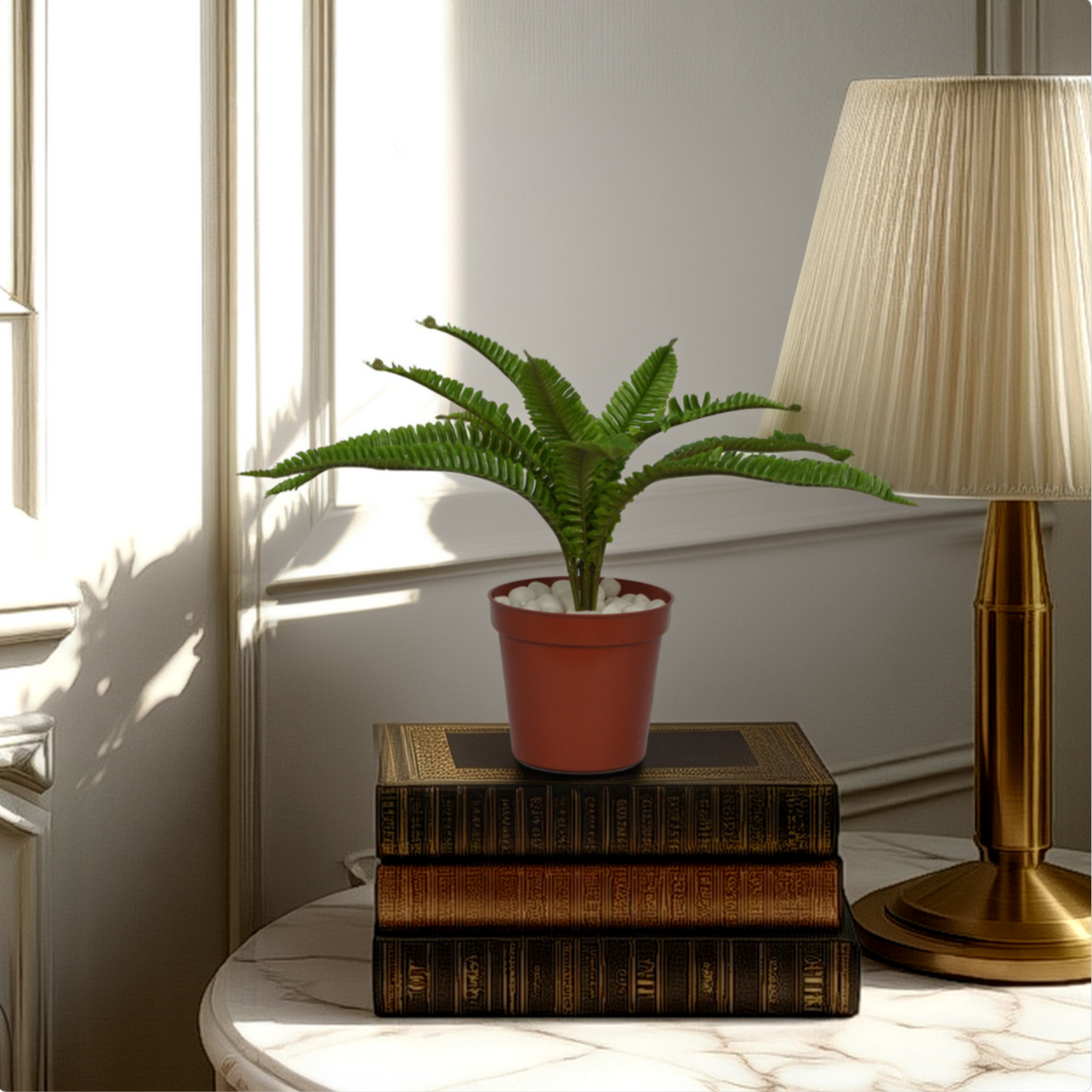 An artificial green fern plant in a brown pot, placed on a stack of books on a table, with a lamp and a window in the background.