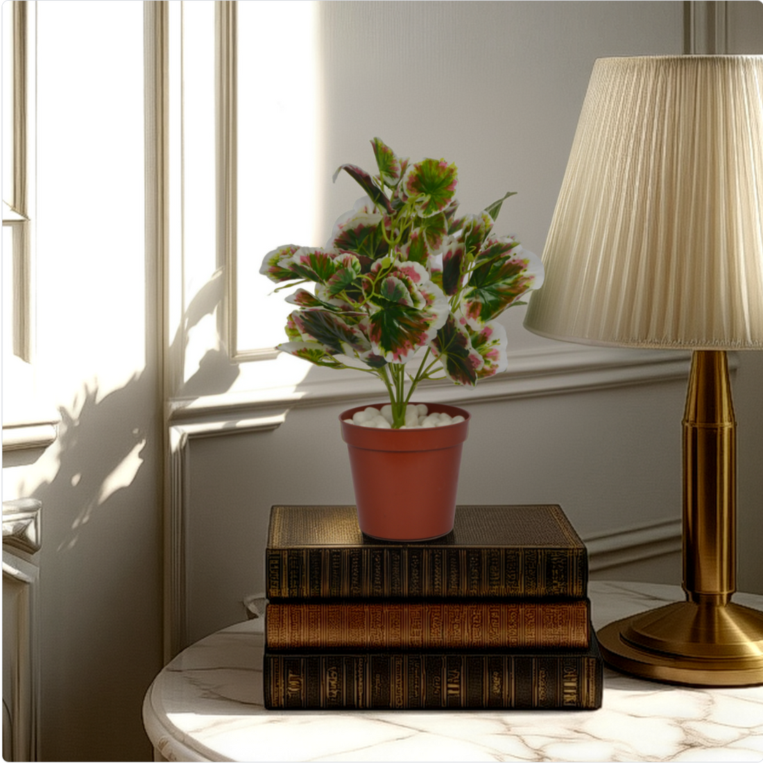 An artificial coleus bush with green, white, and pink leaves in a red pot, placed on a stack of books beside a lamp on a table.