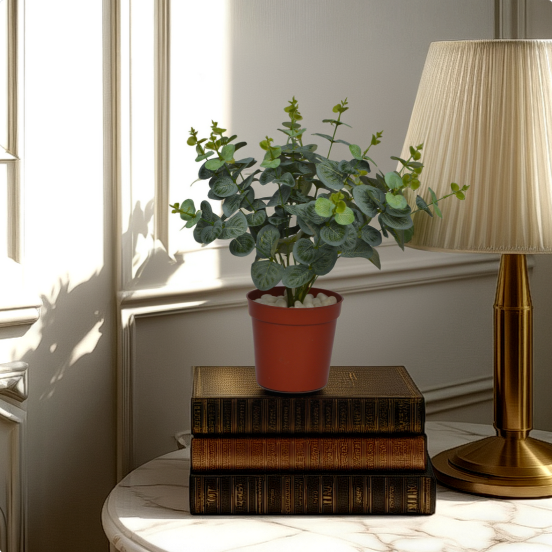 An artificial green carpinteria bush in a red pot, placed on a stack of books on a table, with a lamp and a window in the background.