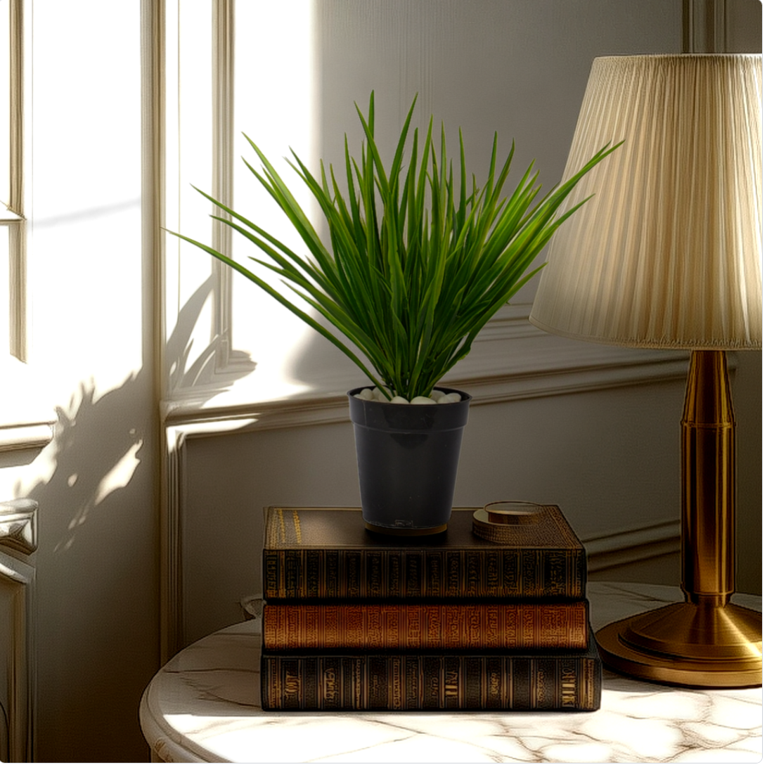 An artificial green grass bush in a navy pot, placed on a stack of old books, next to a lamp, on a white surface, with a window in the background.
