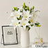 White lilies in a vase on a table with a photo frame and small glass container in the background.